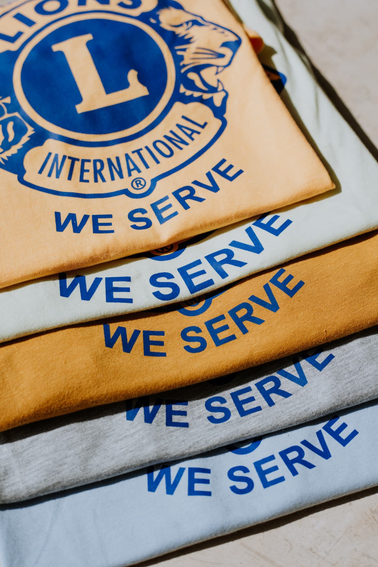 Stack of Lions Club International 'We Serve' printed t-shirts in gold, cream, gray and light blue